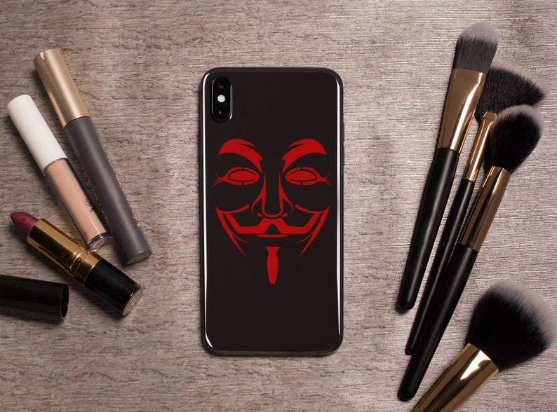 May include: A black phone case with a red Anonymous mask graphic. The phone case is on a wooden surface with makeup brushes.
