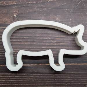 May include: A white, pig-shaped cookie cutter. The cutter is made of a solid, opaque material and has a simple outline of a pig. The cutter is designed for baking and crafting. The background is a dark wood surface.