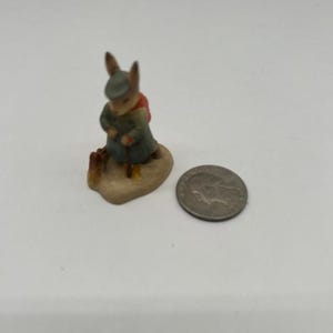 May include: A small figurine of a rabbit wearing a green coat, red scarf, and hat, holding a walking stick. The rabbit is standing on a small, tan base. A silver coin is next to the figurine.