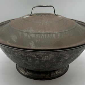 May include: An antique metal cooking pot with a lid. The pot is a dark, weathered gray color with a matching lid and two curved handles. The lid has a small handle on top.