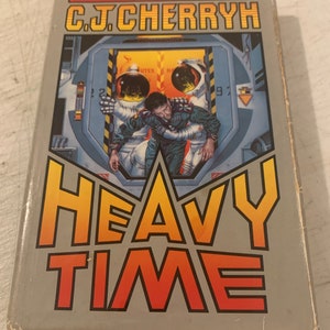 May include: A paperback book cover with a gray background and the title "HEAVY TIME" in large, orange and yellow letters. The author's name, "C.J. CHERRYH", is printed in black above the title. The cover features an illustration of two astronauts in a futuristic space station.