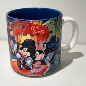 May include: A white ceramic mug with a blue rim and a colourful image of Mickey Mouse and Minnie Mouse in front of a building that says "Club Daisy".