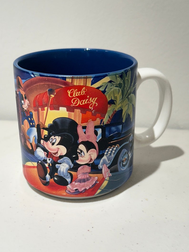 May include: A white ceramic mug with a blue rim and a colourful image of Mickey Mouse and Minnie Mouse in front of a building that says "Club Daisy".