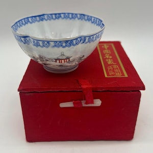 Puede incluir: Un cuenco de porcelana blanco y azul con borde festoneado, con una escena de paisaje. El cuenco está sobre una caja roja con caracteres chinos. La caja tiene un cierre blanco.
