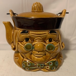 Könnte beinhalten: Eine Vintage-Teekanne aus Keramik in Form eines lächelnden asiatischen Männerkopfes. Die Teekanne ist hauptsächlich beige mit braunen und türkisfarbenen Akzenten. Sie hat einen Deckel, einen Ausguss und einen Griff und ist in einem skurrilen, cartoonartigen Stil gestaltet.
