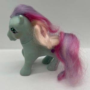 May include: A light blue toy pony with a pink, purple, and blonde mane and tail. The pony has a silver tiara and a heart-shaped gem on its side. The eyes are detailed with blue and green accents.