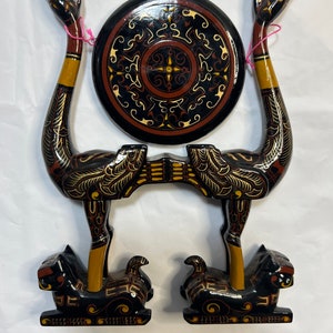 May include: A wooden sculpture of two black and gold birds with outstretched necks, holding a round black and gold painted disc. The birds are standing on black and gold painted animal figures.