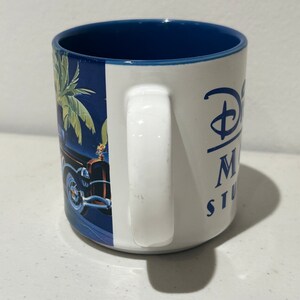 May include: White ceramic mug with a blue rim and a blue and green palm tree design. The mug has a white handle and the text "Disney's Hollywood Studios" in blue lettering.