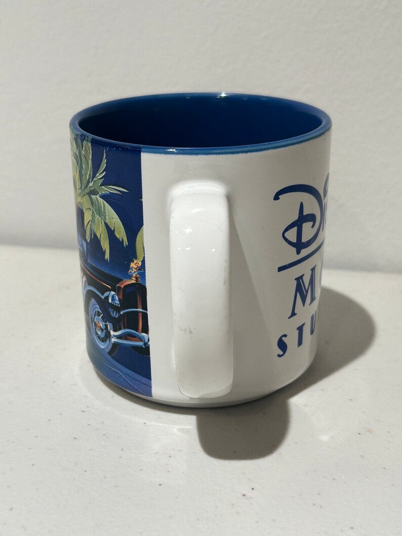 May include: White ceramic mug with a blue rim and a blue and green palm tree design. The mug has a white handle and the text "Disney's Hollywood Studios" in blue lettering.
