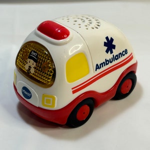 May include: A white toy ambulance with red and yellow accents. The ambulance has a red light on top, a yellow light on the side, and a blue star of life symbol on the side. The words "Ambulance" and "EMT" are printed on the side of the toy. The toy is made by VTech.