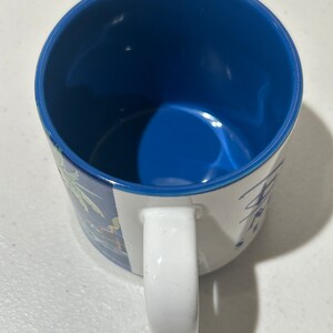 May include: A white ceramic mug with a blue interior and a handle. The mug has a colourful design on the outside.