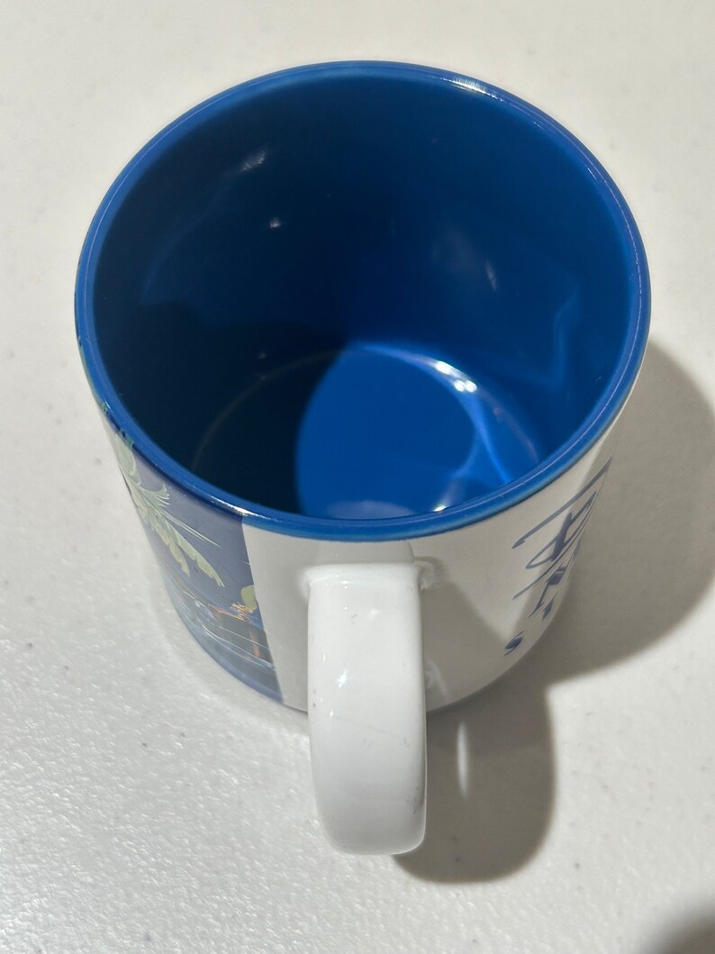 May include: A white ceramic mug with a blue interior and a handle. The mug has a colourful design on the outside.