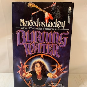 May include: A paperback book cover with a purple and white title "Burning Water" by Mercedes Lackey, author of The Heralds of Valdemar series. The cover features a woman with long dark hair, wearing a black shirt and jeans, with her hands outstretched in front of her. The background is a dark purple with a glowing pentagram.