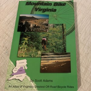 May include: A green book cover with the title "Mountain Bike Virginia" and the subtitle "An Atlas of Virginia's Greatest Off-Road Bicycle Rides" by Scott Adams. The cover features a map of Virginia, a mountain scene, a forest scene, and a close-up of a mountain bike wheel.
