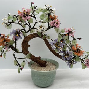 May include: A decorative bonsai tree with a brown trunk and branches, and colorful glass flowers in shades of pink, orange, white, and purple. The tree is planted in a light green ceramic pot filled with small white stones.