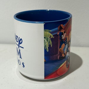 May include: A white ceramic mug with a blue rim. The mug features a graphic of Mickey Mouse and Goofy on the side. The text "Disney MGM Studios" is printed on the mug.