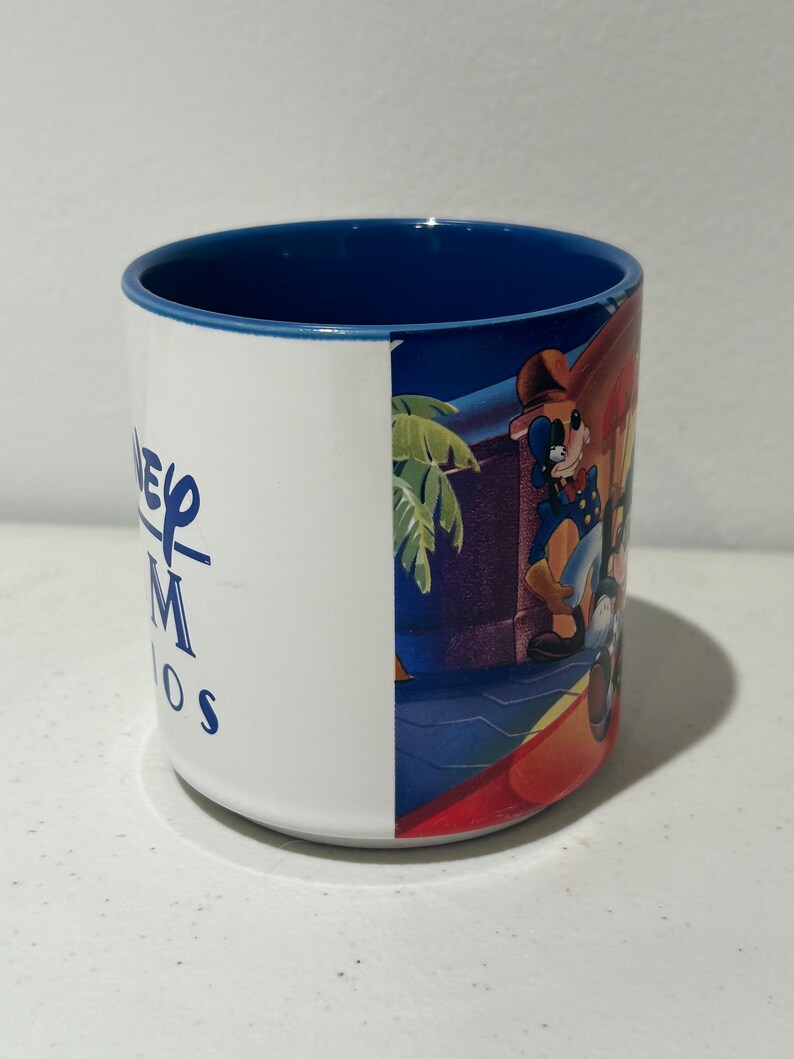 May include: A white ceramic mug with a blue rim. The mug features a graphic of Mickey Mouse and Goofy on the side. The text "Disney MGM Studios" is printed on the mug.