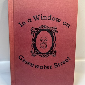 May include: A hardcover book with a textured, dusty rose cover. The title "In a Window on Greenwater Street" is printed in black, along with a decorative frame and a small dog illustration.