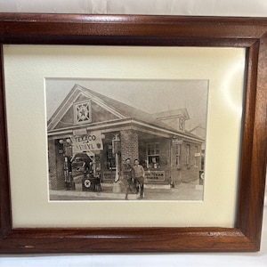 May include: A black and white photograph of a petrol station with two men standing in front. The petrol station has a sign that reads "Texaco Ethyl".