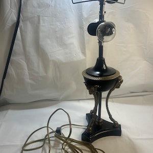 May include: A black and gold table lamp with a globe-shaped shade and a decorative base with three legs. The lamp has a cord with a plug.