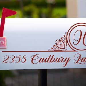 Pode incluir: Caixa de correio branca com uma bandeira vermelha, uma borda decorativa vermelha e um monograma vermelho com a letra "H" em um círculo. O endereço "2358 Cadbury Lane" está escrito em itálico vermelho.
