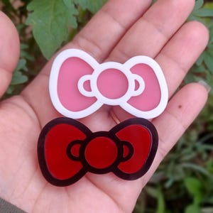 May include: Two plastic bow-shaped hair clips. The top clip is white with a pink bow. The bottom clip is black with a red bow.
