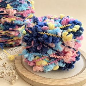 May include: Close-up of two spools of colorful yarn. The yarn is a mix of blue, pink, yellow, and light blue, with a textured, pom-pom-like appearance. One spool is in focus, sitting on a wooden coaster, while the other is slightly blurred in the background.