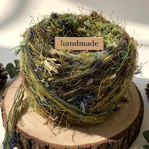 May include: A ball of textured yarn in shades of olive green, brown, and muted blue, featuring a rectangular wooden tag with the word handmade printed in black. The yarn has a loose, fibrous construction with varying thicknesses and loose strands.