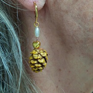 Peut inclure: Boucle d'oreille dorée avec un design pendant. La boucle d'oreille comprend une perle blanche, une perle dorée en forme de cœur et un charm en forme de pomme de pin jaune. La boucle d'oreille est portée à l'oreille d'une personne.