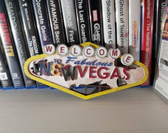 Może przedstawiać: Żółto-czerwony znak z napisem "Welcome to Fabulous Las Vegas" białymi literami. Znak jest w stylu vintage i ma zużyty wygląd. Znak stoi na półce z grami wideo za nim.