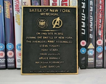May include: A bronze plaque commemorating the Battle of New York, a scene from the Avengers movie. The plaque features the New York City seal and the Avengers logo. It lists the names of the Avengers who assembled during the battle: Steve Rogers, Tony Stark, Thor Odinson, Bruce Banner, Natasha Romanoff, and Clint Barton. The plaque is mounted on a wall with a shelf of DVDs behind it.