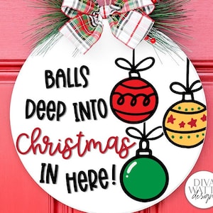 May include: A white round door hanger with the text "Balls deep into Christmas in here!" in black and red lettering. There are three cartoon Christmas ornaments hanging from the top of the sign. The ornaments are red, yellow, and green.