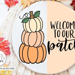 Könnte beinhalten: Ein rundes Holzschild mit einem Stapel von drei Kürbissen und dem Text "Willkommen auf unserem Feld" in schwarzer Schrift. Die Kürbisse sind orange, pfirsichfarben und cremefarben.