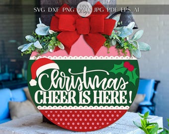 Pode incluir: Uma placa de Natal redonda com um laço vermelho, vegetação e o texto "Christmas CHEER IS HERE!". A placa apresenta um gorro de Papai Noel, azevinho e um padrão de bolinhas vermelhas. A placa tem aproximadamente 30 cm de diâmetro.