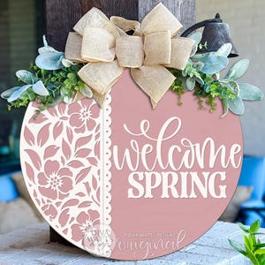May include: A circular pink wooden sign with the words "Welcome Spring" in white script. The sign has a floral cut-out design on the left, a burlap bow, and greenery. A small bell hangs above the sign.
