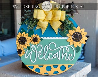 May include: A round wooden welcome sign with a blue and green background. The sign features a yellow bow, green leaves, and sunflowers. The word "Welcome" is written in white cursive lettering.