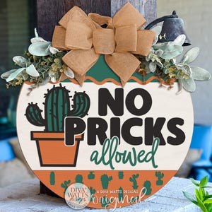 May include: Round wooden sign with a cactus illustration and the text "NO PRICKS allowed". The sign features a tan bow, green and white foliage, and a decorative border with cactus silhouettes. The sign is a decorative item.