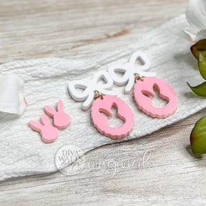 May include: Pink Easter bunny earrings and matching bunny stud earrings with a white bow accent. The earrings are on a white cloth with white flowers and green leaves in the background. The text "Diva Walls Original" is visible.