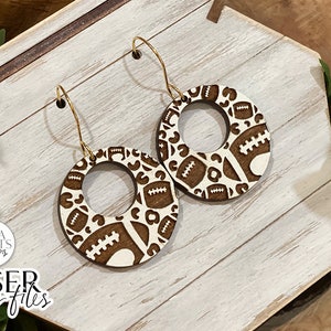 May include: A pair of wooden earrings with a brown and white football pattern. The earrings are round with a hole in the centre and have gold hooks.