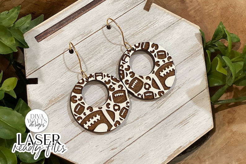 May include: A pair of wooden earrings with a brown and white football pattern. The earrings are round with a hole in the centre and have gold hooks.