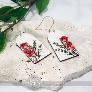 May include: A pair of white wooden earrings with a floral design. Each earring features a red rose and green leaves. The earrings have gold-colored hooks and are displayed on a textured, light-colored surface.