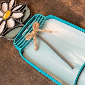May include: A teal blue mason jar with a white wash finish and a brown twine bow. A white daisy with a yellow center is attached to the jar.