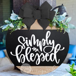 Puede incluir: Un letrero redondo de madera negra con las palabras "Simply Blessed" en escritura blanca. Decorado con un gran lazo negro, vegetación y una pequeña campana. Un objeto decorativo.