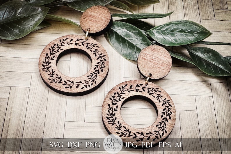 May include: A pair of wooden earrings with a circle design featuring a leafy wreath. The earrings are brown and have a gold-colored metal hook.
