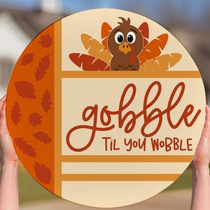 May include: A round wooden sign with a cartoon turkey and the words "gobble til you wobble" in a decorative font. The sign has orange and tan accents and is held by two hands.