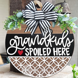 May include: A round wooden sign with the text "grandkids SPOILED HERE" in white script, a red heart, and a decorative white pattern. The sign is adorned with a black and white striped bow and greenery.