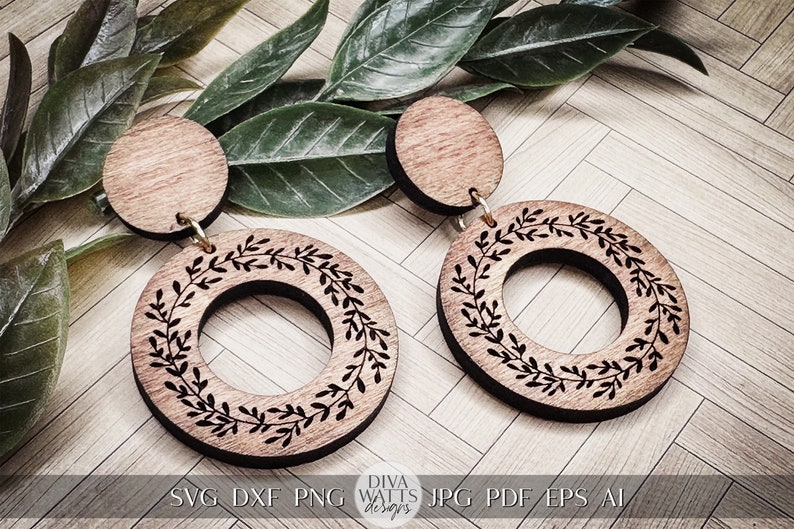 May include: A pair of wooden earrings with a circular design featuring a wreath of leaves. The earrings are brown with a natural wood finish and have a simple, elegant style.