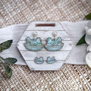 May include: A set of earrings and matching studs featuring a blue bird in a nest design. The earrings have pearl accents and hang from gold-colored hooks. The jewelry is displayed on a white and gray wooden hexagon-shaped stand.