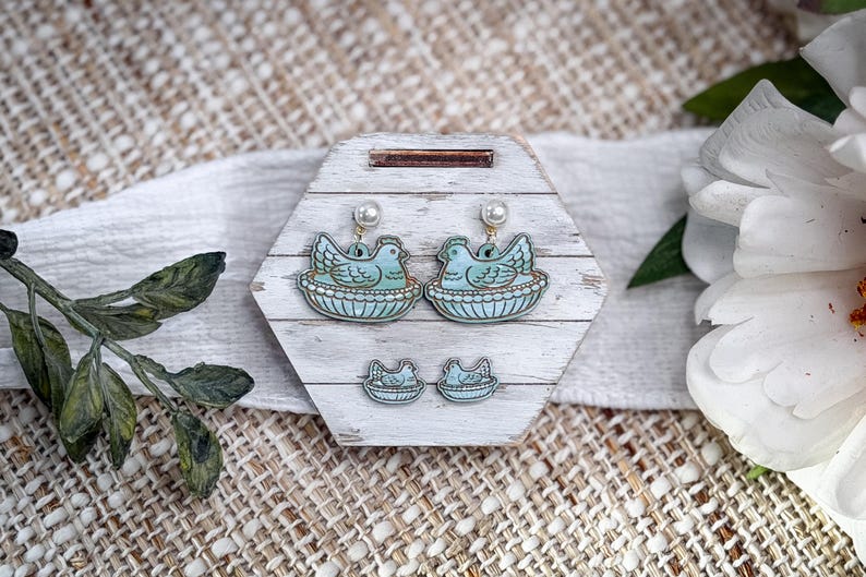 May include: A set of earrings and matching studs featuring a blue bird in a nest design. The earrings have pearl accents and hang from gold-colored hooks. The jewelry is displayed on a white and gray wooden hexagon-shaped stand.