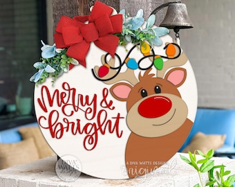 Puede incluir: Un letrero navideño redondo de madera blanca con un diseño de reno de dibujos animados. El letrero presenta el texto "Merry & Bright" en escritura roja, un lazo rojo y una cadena de luces de colores. Una pequeña campana cuelga de la parte superior.